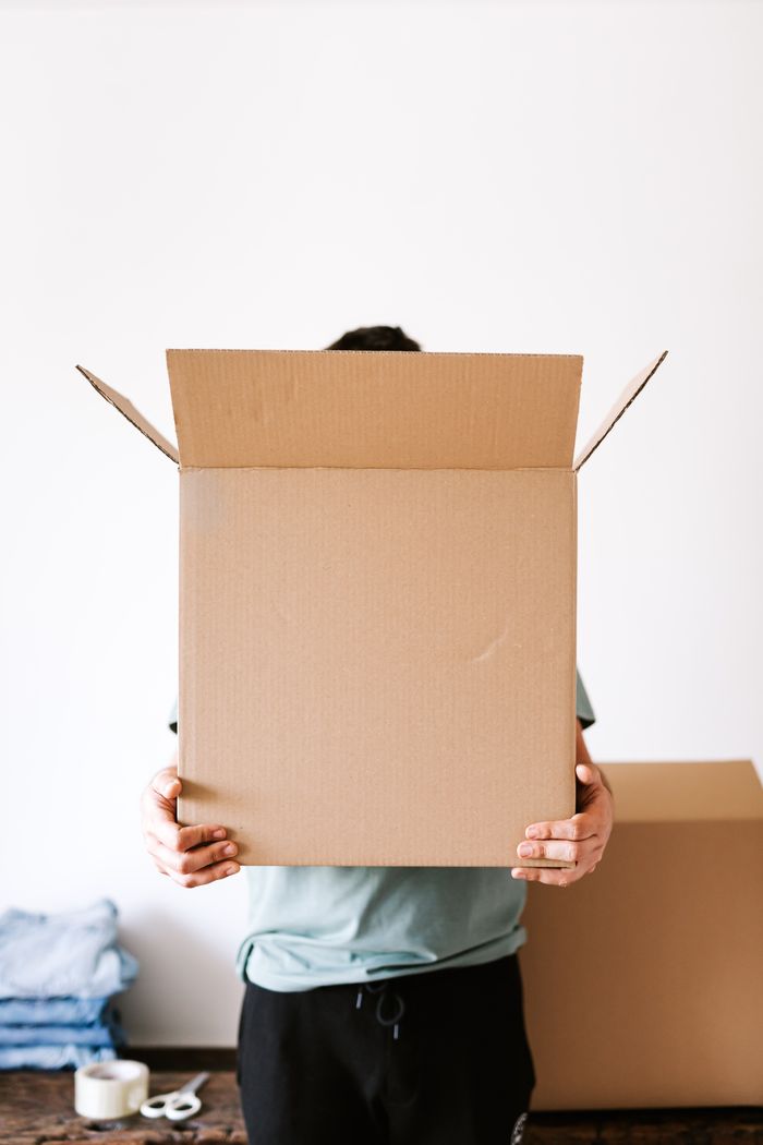 why-choose-us-02 Anonymous man carrying opened carton box before packing belongings and relocation while standing against white wall and stacked clothes